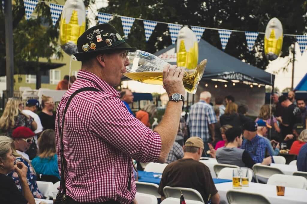 A man wearing traditional Bavarian attire drinks from a glass boot